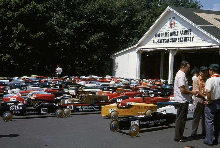 Many soap box derby cars outside a building with a sign reading "Home of the World Famous Soap Box Derby"